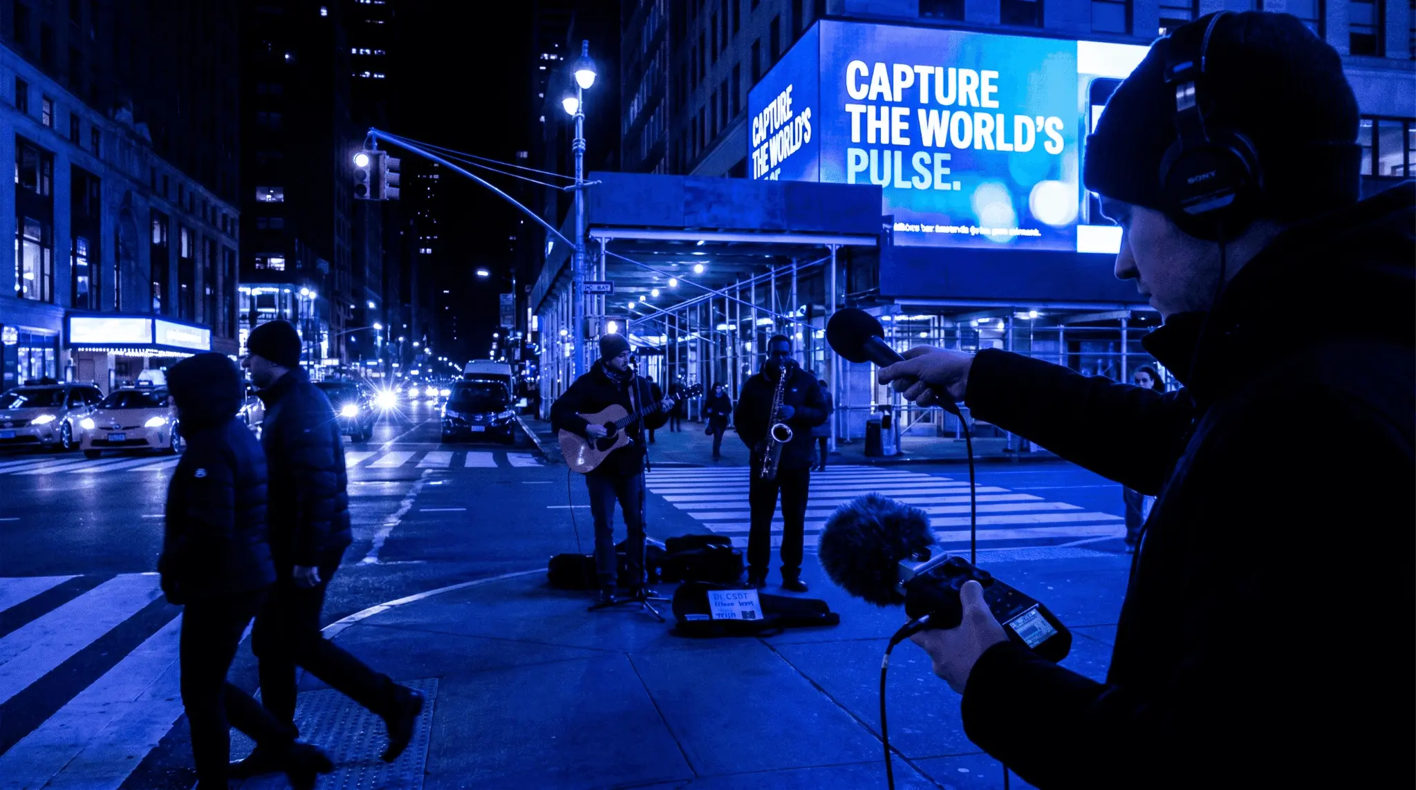 Producer recording street musicians and city ambience with a handheld recorder and microphone for field recording and found sound sampling.