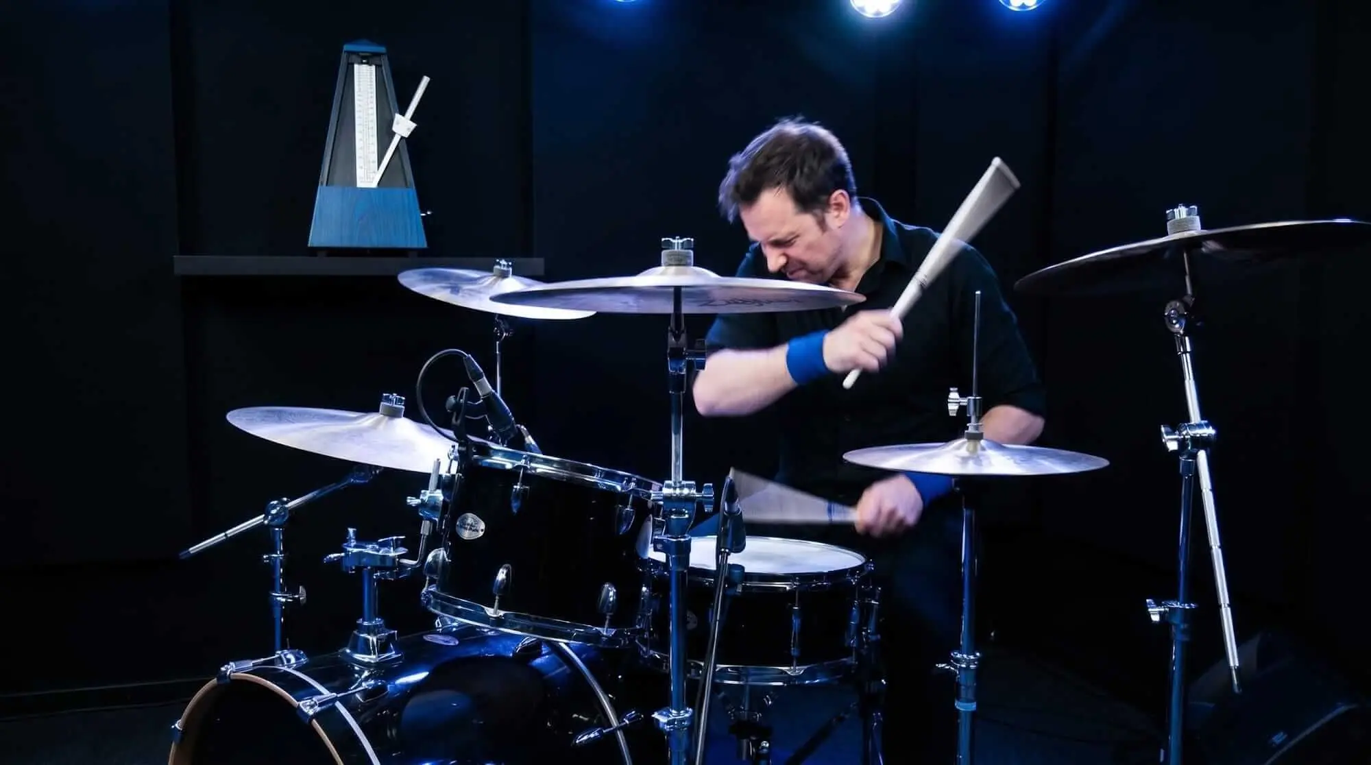 Baterista actuando en una batería acústica junto a un metrónomo en un estudio oscuro.